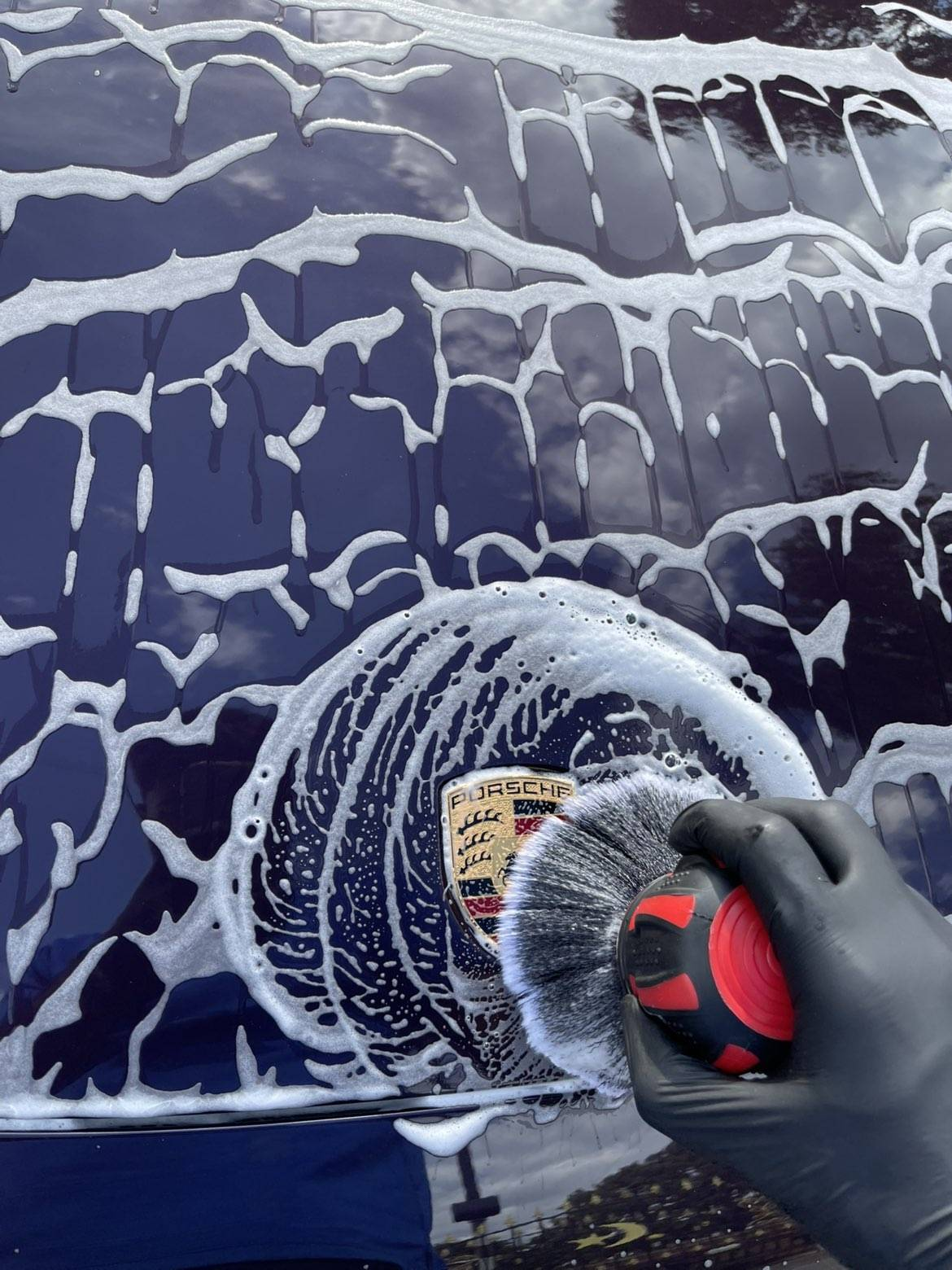 Detailer applying snow foam and detailing brush to a navy Porsche badge